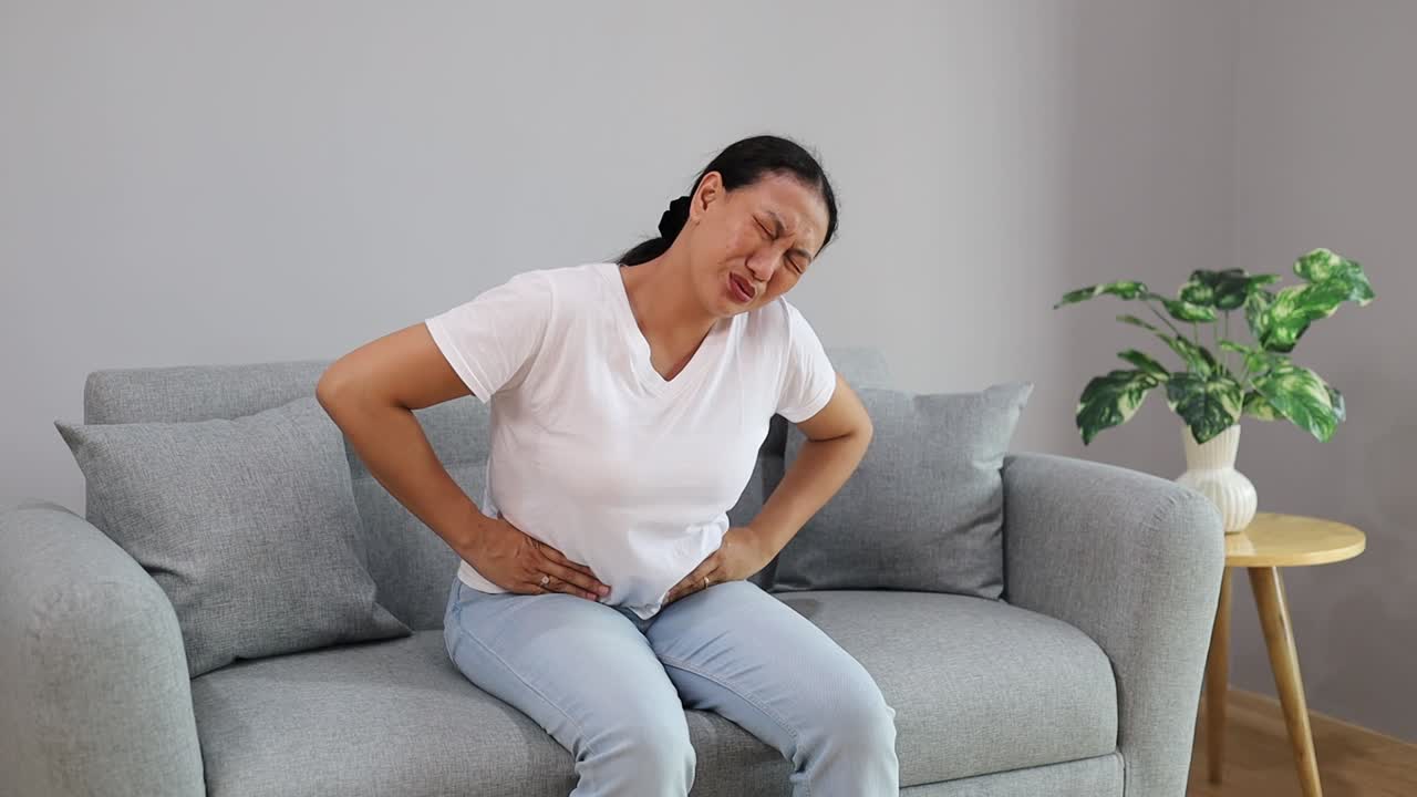 Asian adult woman sitting on couch is experiencing intense abdominal pain. She is holding her belly and grimacing in discomfort