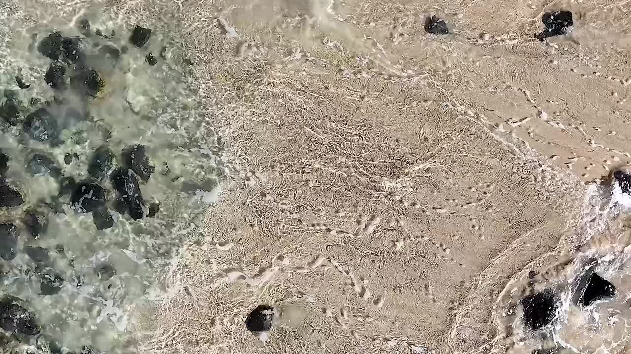 Close-up aerial footage of waves crashing over a rocky shoreline with clear, turquoise waters.