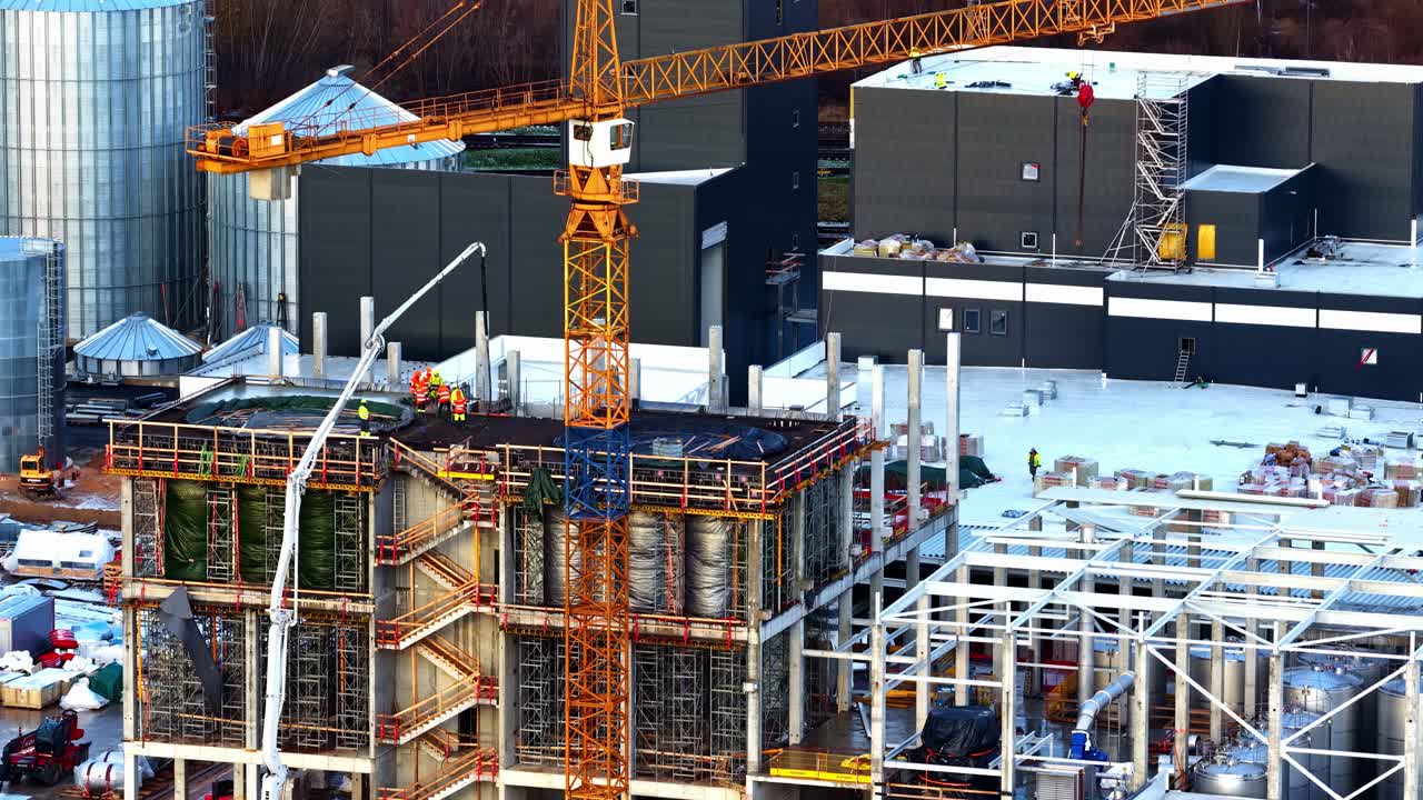 Drone close-up of crane and workers on snowy industrial construction site with concrete columns, steel framework, and scaffolding during winter building project