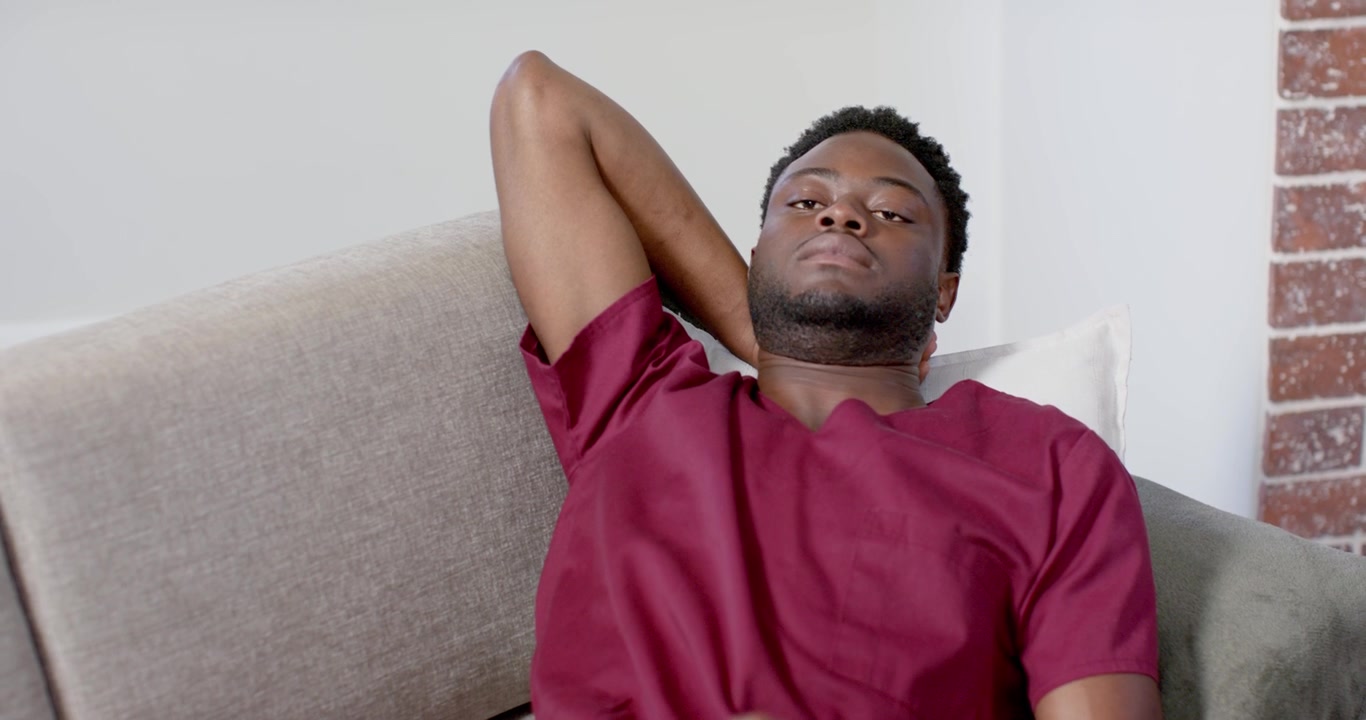 Afro-Amerikaanse man wordt wakker op de bank in bordeauxrode ziekenhuiskleding, tilt hoofd op en controleert smartwatch.