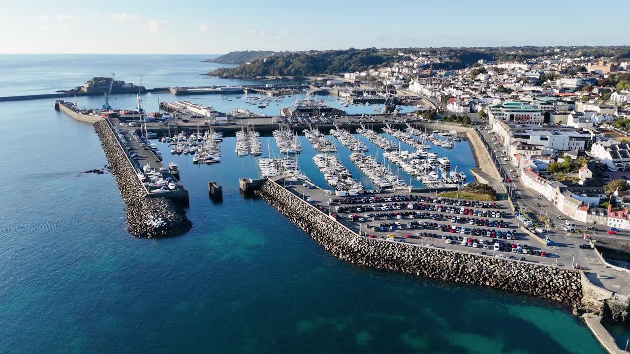 Volo drone all'indietro su St Peter Port, Guernsey, sopra la QEII Marina e l'angolo Salerie, con lo sfondo del Castello Cornet e del porto, con uno splendido mare calmo e limpido di un blu profondo sotto il sole del tardo pomeriggio