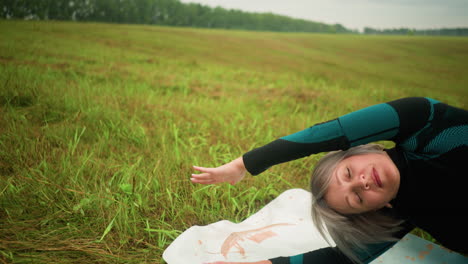 old woman with lying one side on yoga mat practicing side bend pose with arm extended, in a vast grassy field under cloudy skies, surrounded by nature and trees in the distance