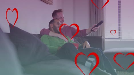 Animation-of-heart-icons-over-diverse-gay-couple-smiling-and-watching-tv