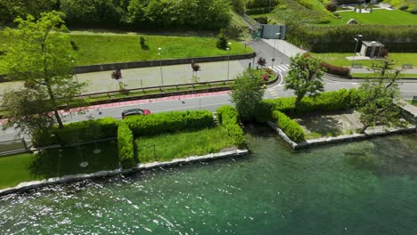 Tracking-aerial-view-of-red-car-on-asphalt-road-next-to-lake-in-town,-Pella,-Italy