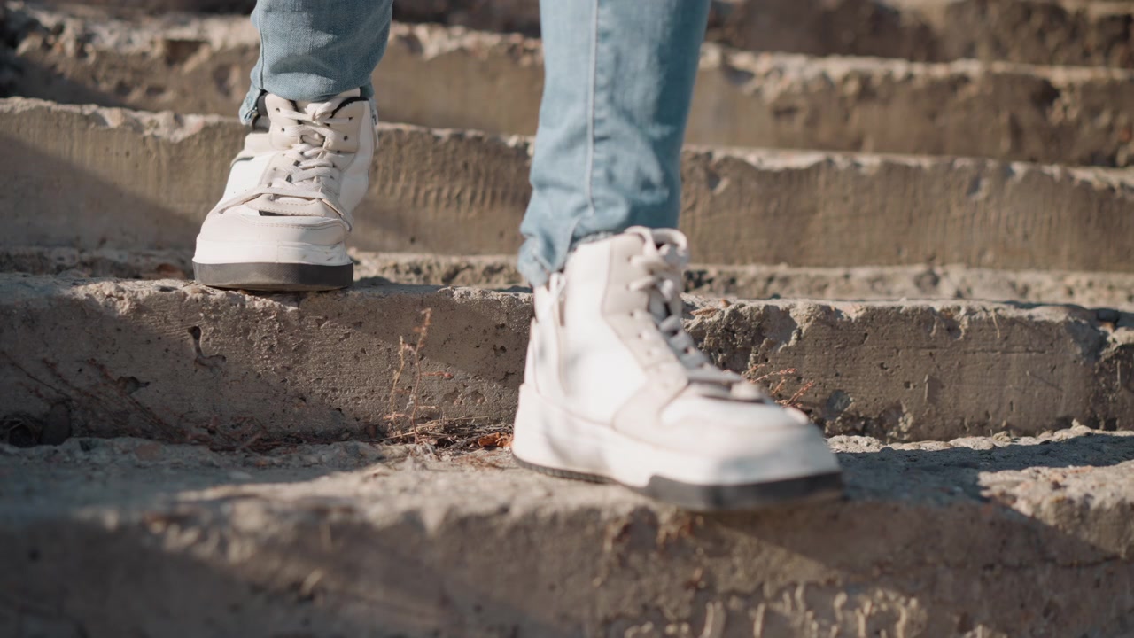 Woman feet in white sneakers and blue jeans ascend concrete steps outdoors. focus remains on movement of her legs capturing texture details like laces or scuffs on shoes against rough stone