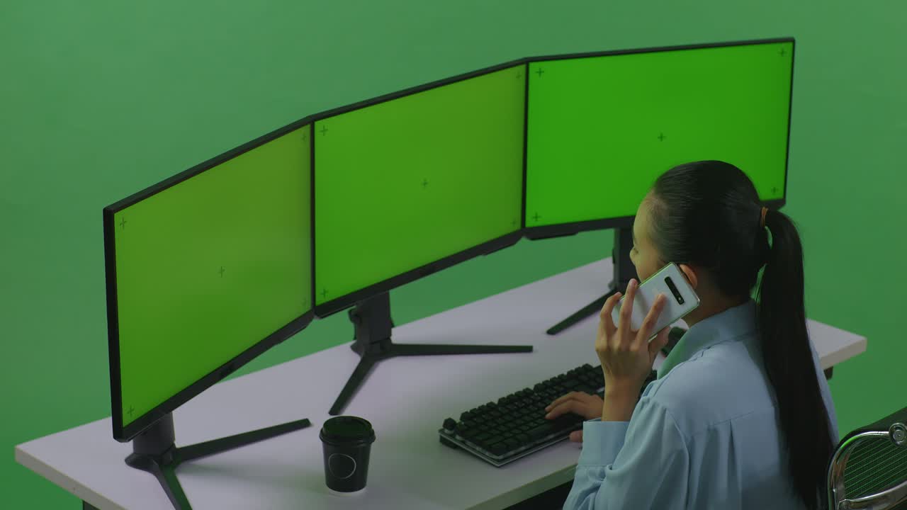 vista lateral de una mujer asiática hablando en un teléfono móvil mientras trabaja con múltiples monitores de computadora simulados en un estudio de pantalla verde