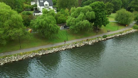 A-female-running-a-long-sida-of-a-small-lake-with-trees-and-a-white-house-in-the-background