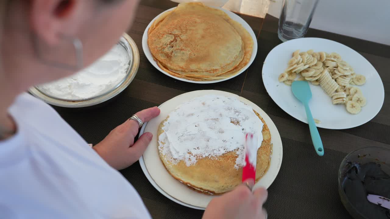 Over the shoulder view of woman spreading whipped cream on a crepe cake using red spatula