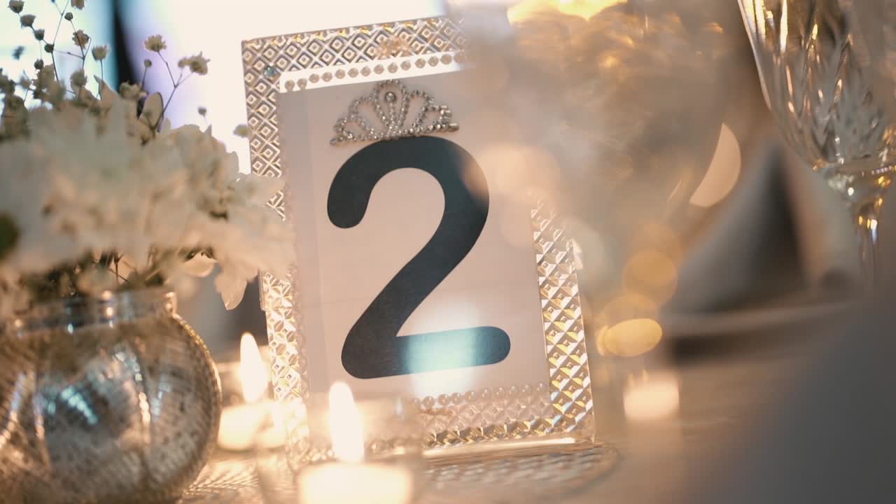 Close-up of table number two displayed on a white decorated table with candles and flowers. Telephoto shot with shallow depth of field, blurred glassware in the background,
