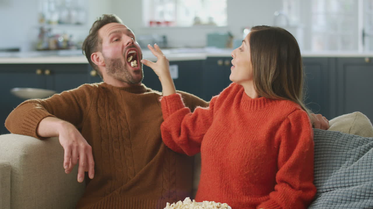Man Catching Popcorn In Mouth As Woman Throws It To Him Sitting On Sofa