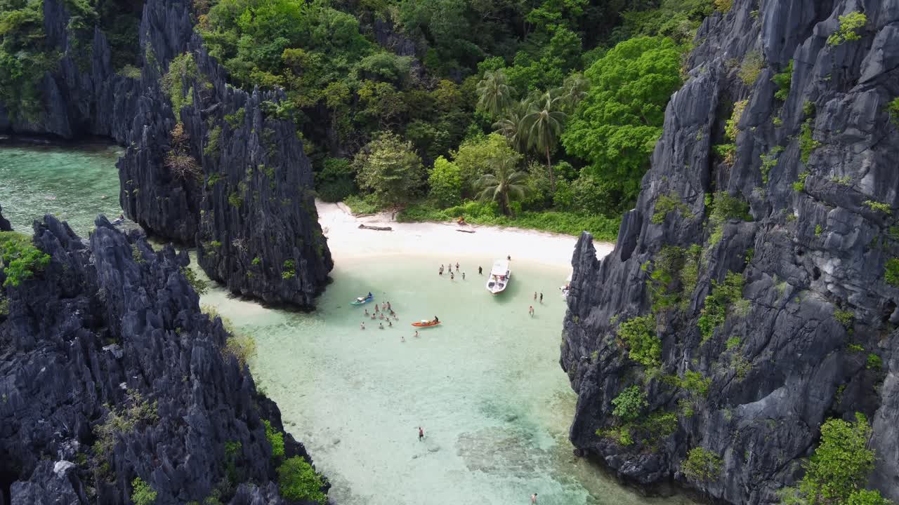 Toma aérea con acercamiento y descenso: Barcos de excursión de isla en isla (Tour C) en la arena blanca de la playa Escondida entre rocas kársticas y selva tropical. Turistas comprando a vendedores en kayak y nadando en la laguna cristalina, El Nido