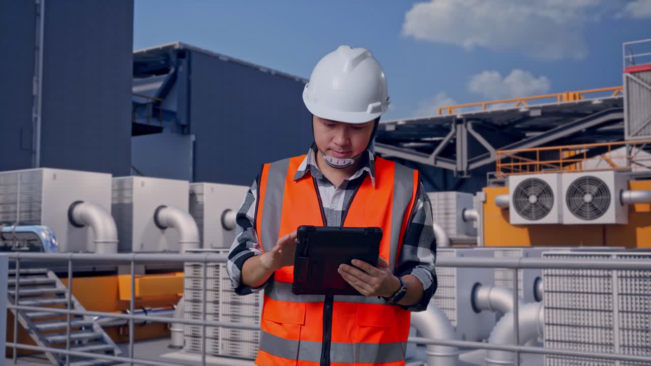 Ingeniero asiático con casco de seguridad trabajando en una tableta mientras está de pie, inspeccionando unidades HVAC en la azotea de un edificio industrial.
