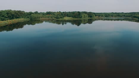 a drone lifts off over a line of trees and flies across a calm scenic waterway surrounded by green trees