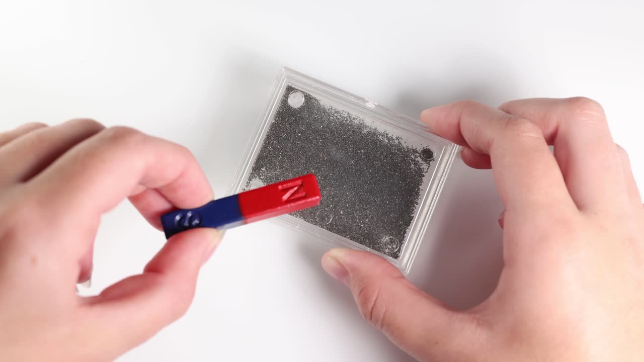 Hands manipulate a magnet over iron filings, illustrating magnetic field patterns on a white background