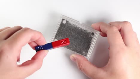 hands manipulate a magnet over iron filings, illustrating magnetic field patterns on a white background