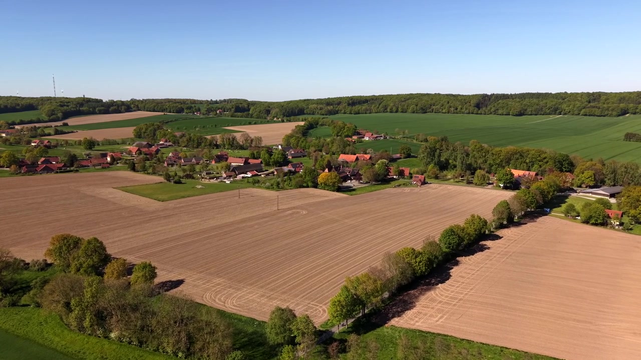 Droge en groene landbouwvelden met boerderijgebouwen en een klein dorp omringd door bomen in de lente. Landelijk platteland en gecultiveerd landbouwlandschap overdag bij zonnig weer. Breed luchtbeeld