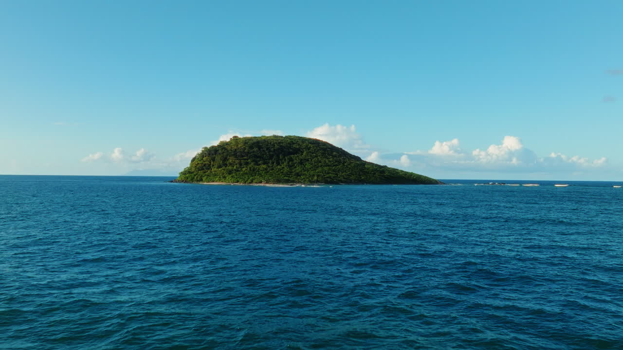 Drone flies forward and rises over the blue ocean toward a lush green island