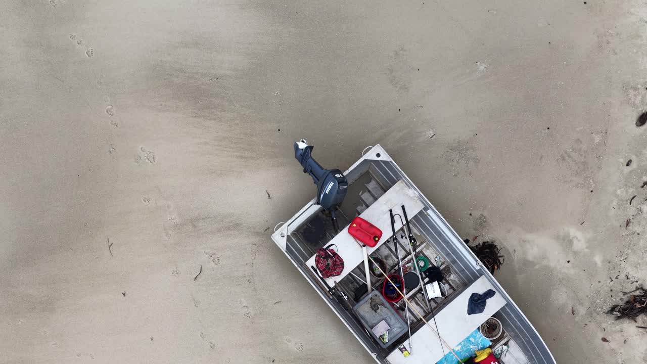 Drone pans over deserted aluminum motorboat with gear on remote, overcast Queensland beach