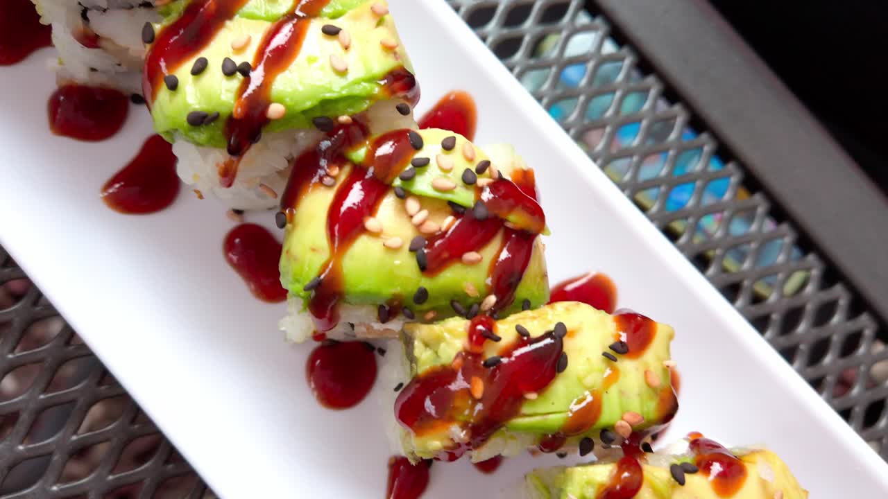 Fresh sushi rolls with avocado, sesame seeds, and sweet sauce served on a white plate