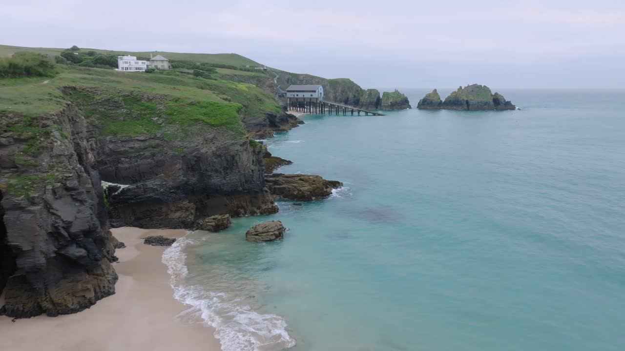 Grassy dark sea cliffs and sandy beach with clear blue seawater with camera flight towards lifeboat station with slipway. Mother Ivey's Bay, Cornwall, UK.