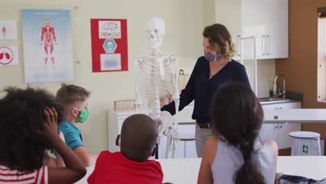 maestra que usa una máscara facial usando un modelo de esqueleto humano para enseñar a los estudiantes en clase