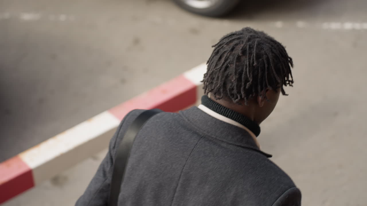 overhead view shows gentleman with dreadlocks in gray coat walking past quiet lane near red white curb barrier, shoulder strap visible, calm afternoon mood, soft focus background suggests solitude