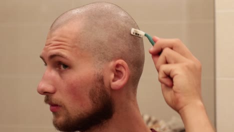 young bald man shaving his own head looking in the mirror with disposable razor, still medium shot