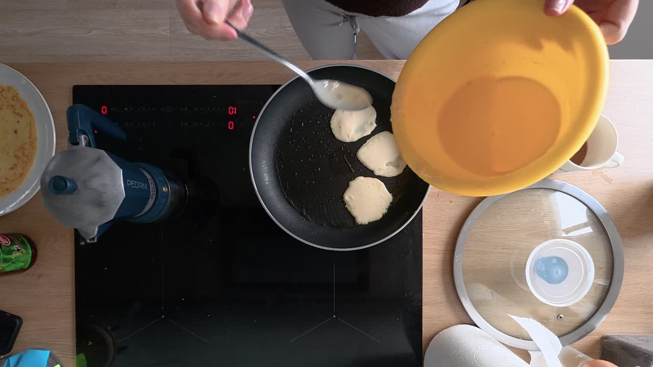 A person carefully adds pancake batter to a pan on a sleek induction cooktop. Warm kitchen light highlights skillful cooking technique with top view composition