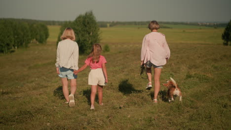 mother and daughter walk closely together, dog owner alongside them as playful dog reaches out for her hand, all enjoying sunny day in vast open field with distant trees in background