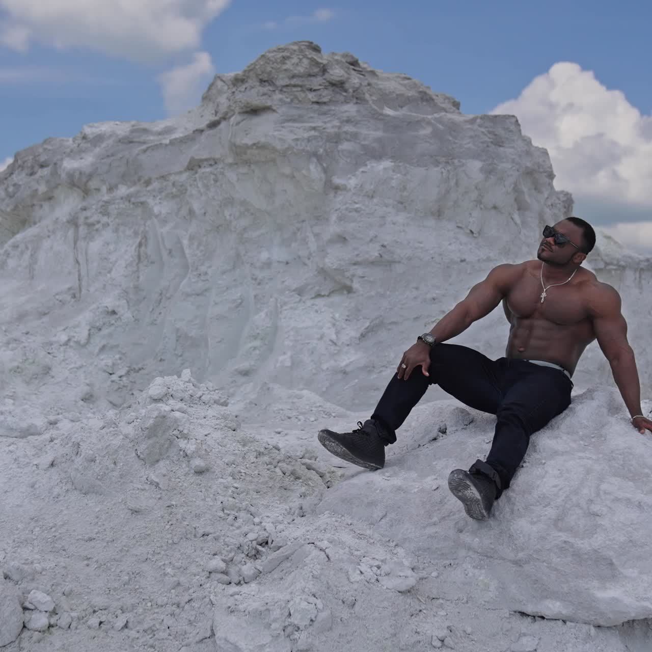 Man with perfect body pumping up muscles and enjoying of the summer. Multiracial man sitting at the sand. Grey rocks background