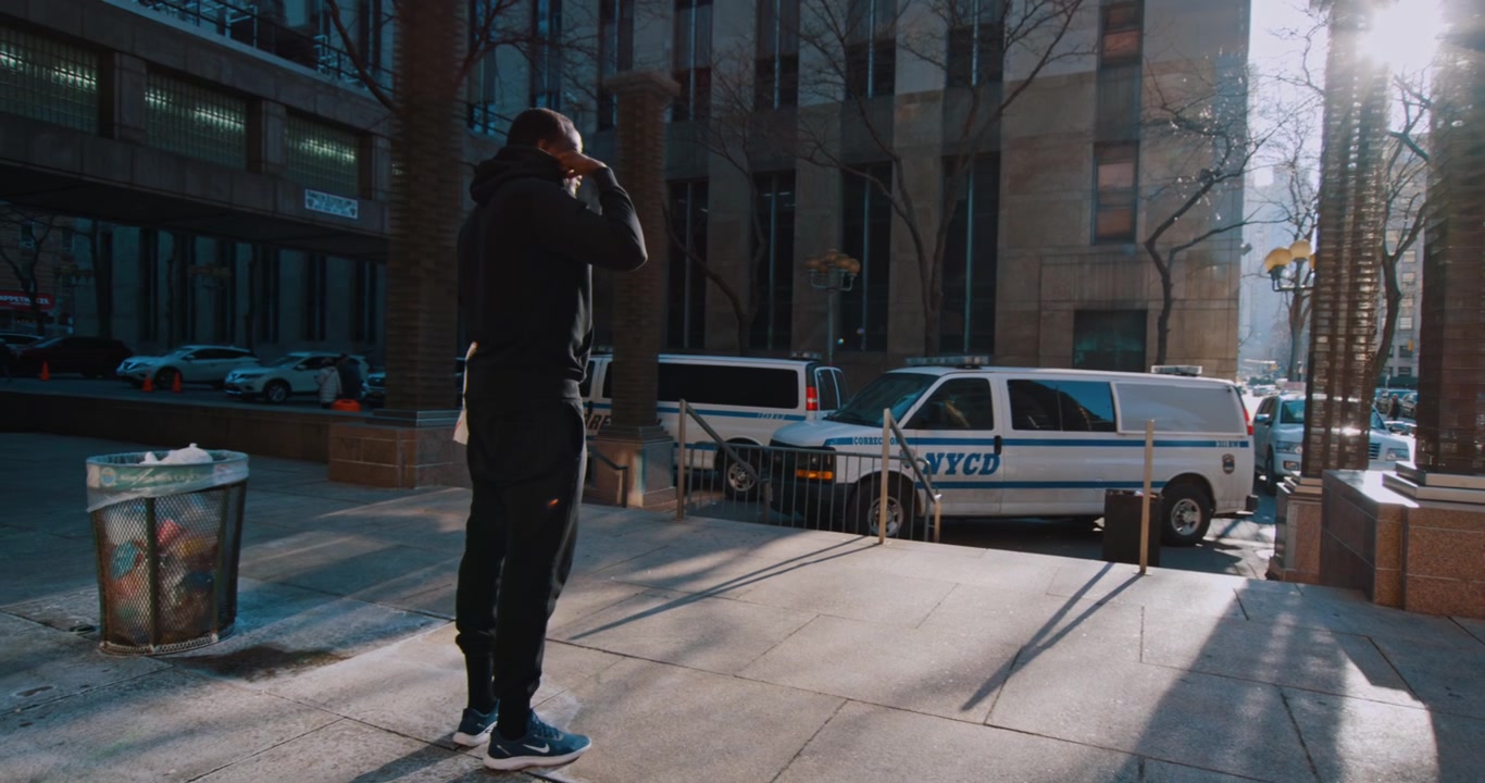 New York, USA, 21 May 2023: Black Man in Black Hoodie Walking on Urban Sidewalk at Dusk. A Black man in a black hoodie and joggers walks in profile along a sidewalk with a stone building in blue light