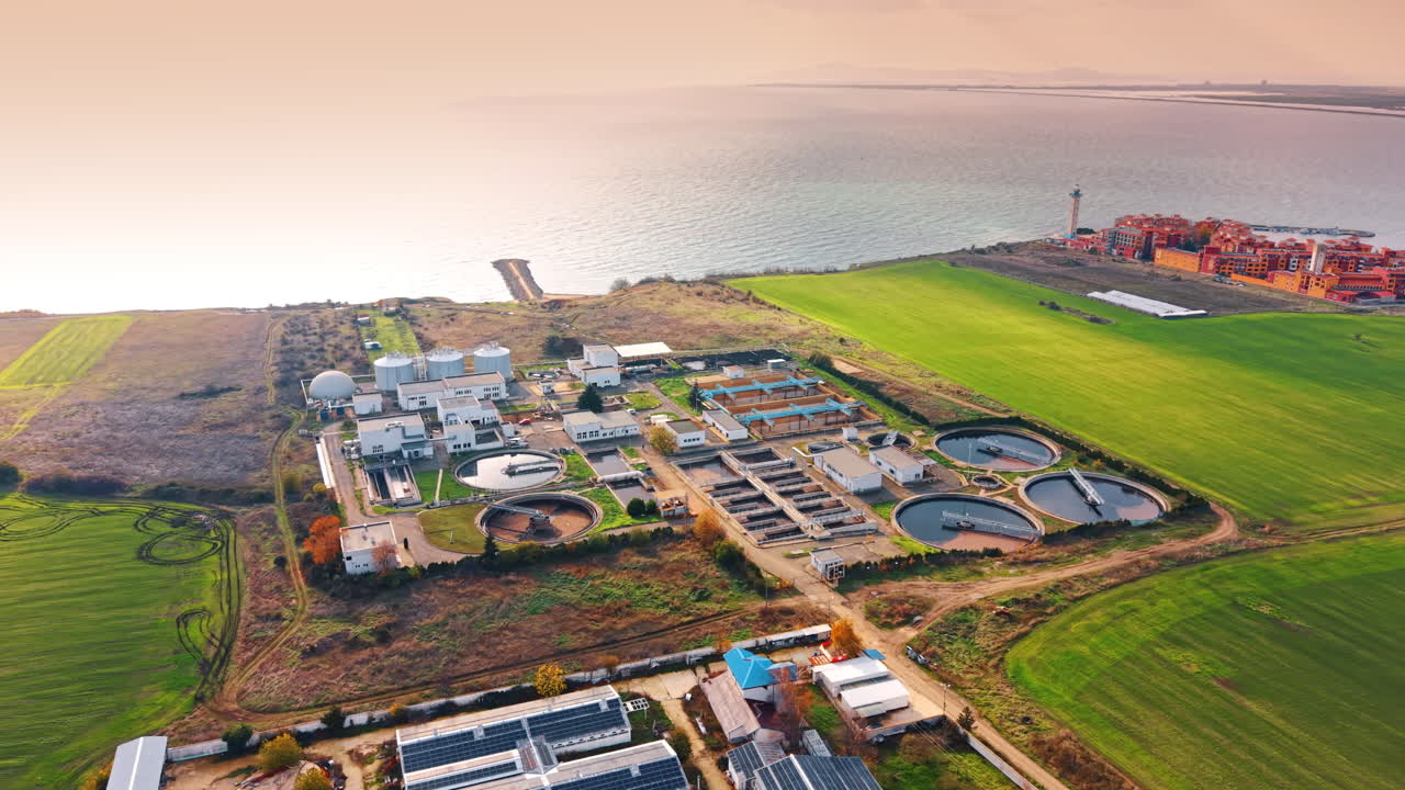 Modern water treatment plant located on the sea shore. A residential area with a lighthouse across the green field at backdrop. Aerial view