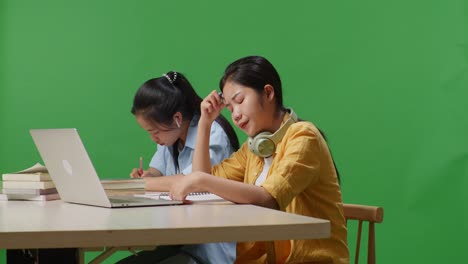 asian woman student is bored while sitting with her friend and writing into the notebook on a table with a laptop in the green screen background classroom