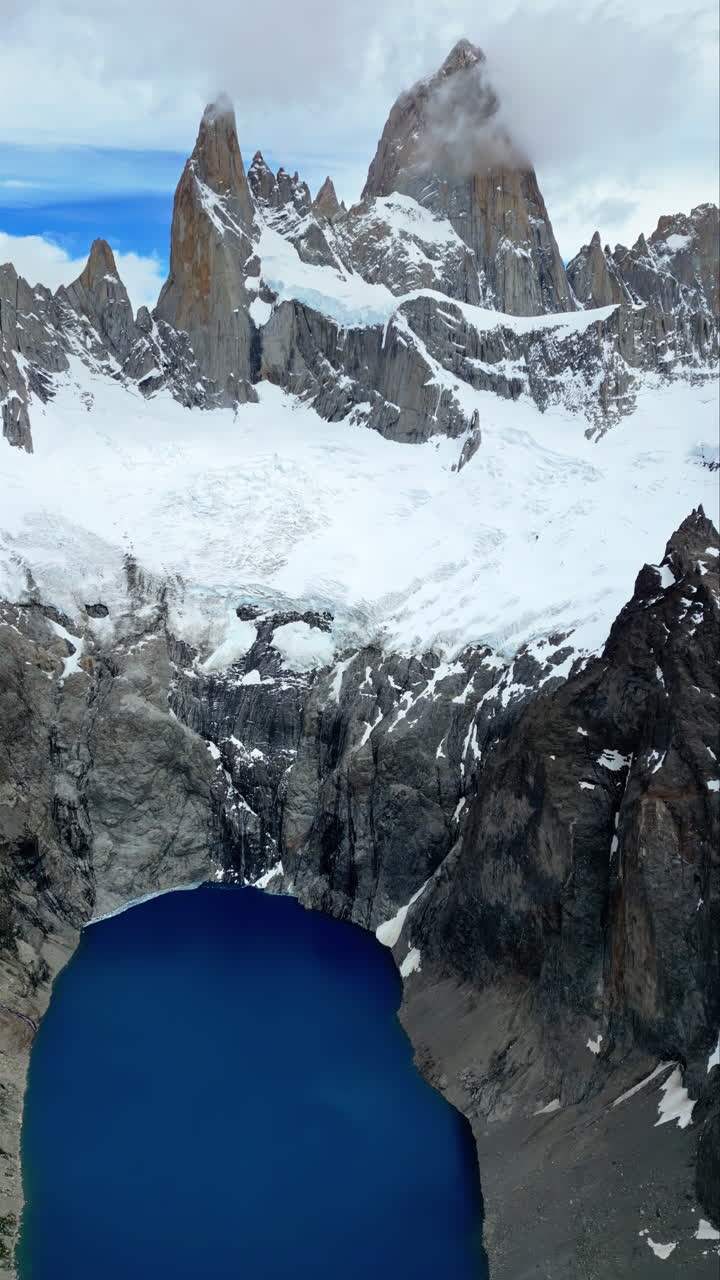 Vista aerea con drone delle maestose cime del Monte Fitz Roy che si ergono sopra un lago glaciale blu intenso, circondato da scogliere rocciose. Verticale