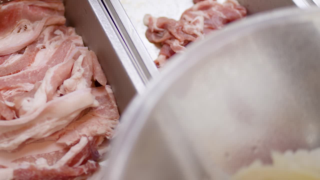 Hand with tongs selecting raw pork belly slices from stainless buffet tray under bright lighting