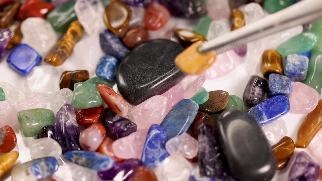 A close-up sequence of tweezers picking a tiger's eye stone from a vibrant collection of chakra stones