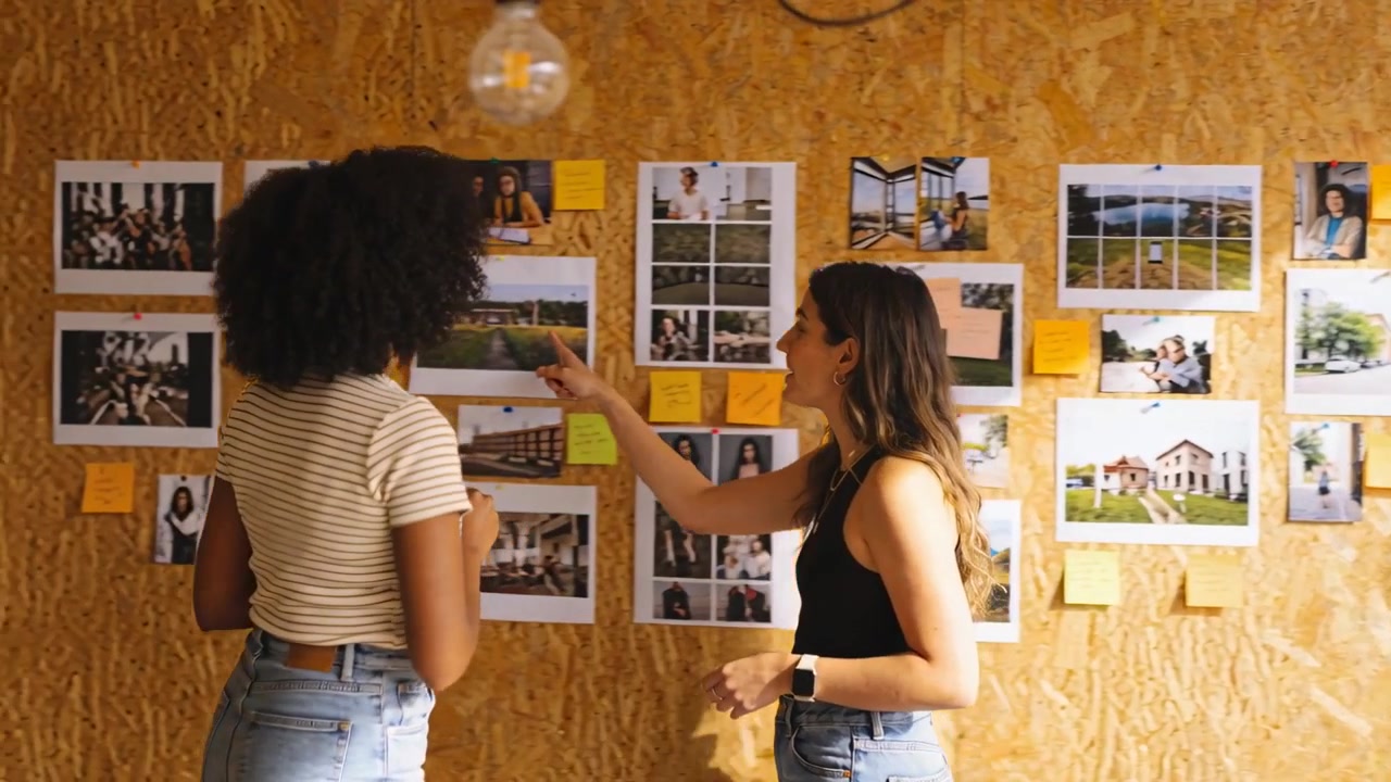 Dos mujeres frente a un panel de contrachapado, organizando impresiones y colocando notas adhesivas para el diseño, con blusas a rayas.