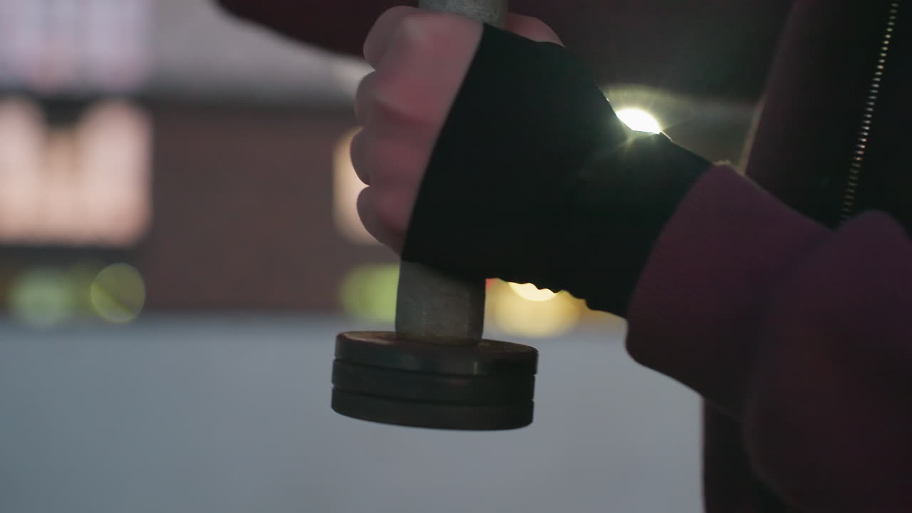 Close hand view of outdoor trainer wearing hoodie and gloves gripping dumbbells, punching air in rapid boxing motion against urban blurred dusk lights background on asphalt court next to fence