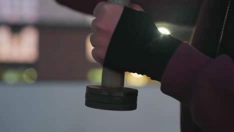 close hand view of outdoor trainer wearing hoodie and gloves gripping dumbbells, punching air in rapid boxing motion against urban blurred dusk lights background on asphalt court next to fence