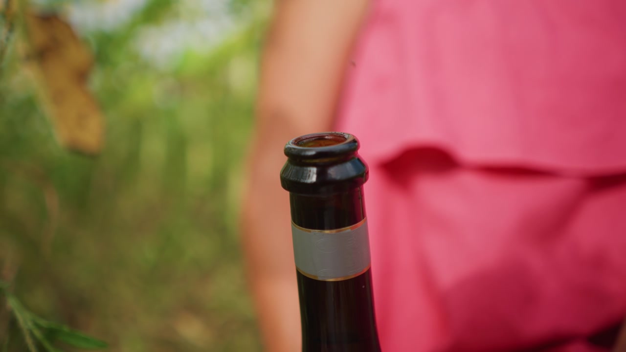 Primo piano di una bottiglia di vetro scuro con etichetta argentata tenuta davanti a una persona sfocata che indossa abiti rosa. Lo sfondo presenta fogliame verde che suggerisce luce naturale esterna e una messa a fuoco morbida.