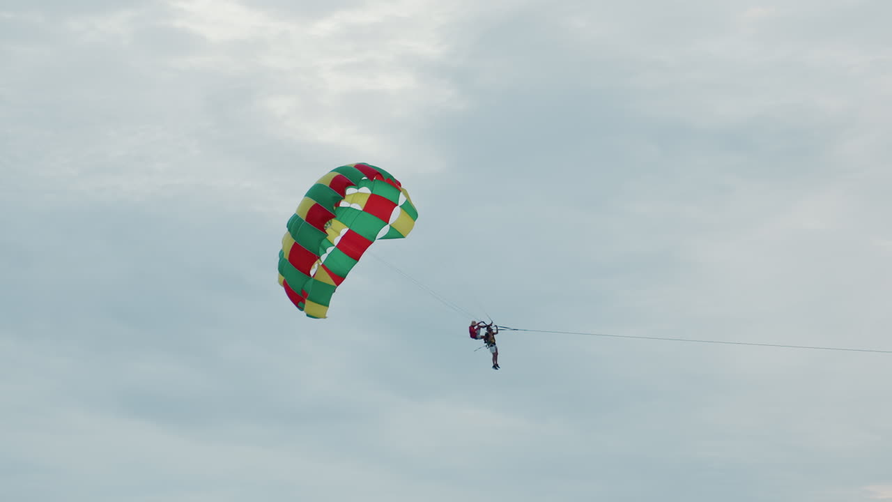 Parasailer sopra l'orizzonte dell'isola all'alba, cielo calmo, baldacchino colorato che scivola sulla silhouette di un'isola distante, movimento lento e rilassato, passeggero legato sospeso sopra l'acqua costiera, brezza leggera, nuvola soffice.