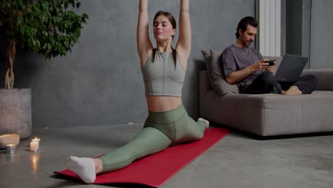 una confiada chica morena en una camiseta gris hecha de pantalones verdes se sienta en las divisiones en una alfombra deportiva roja y hace yoga estiramiento y deportes cerca de ella su novio que está haciendo trabajo usando su portátil en casa en un apartamento moderno cerca de las velas quemadas de una gran planta de interior