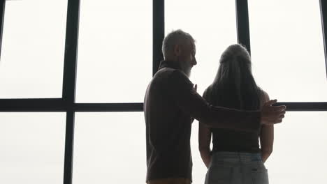 Back-View-Of-Middle-Aged-Man-Embracing-His-Sad-Wife-And-Trying-To-Comfort-Her-While-Standing-Together-At-Window-At-Home