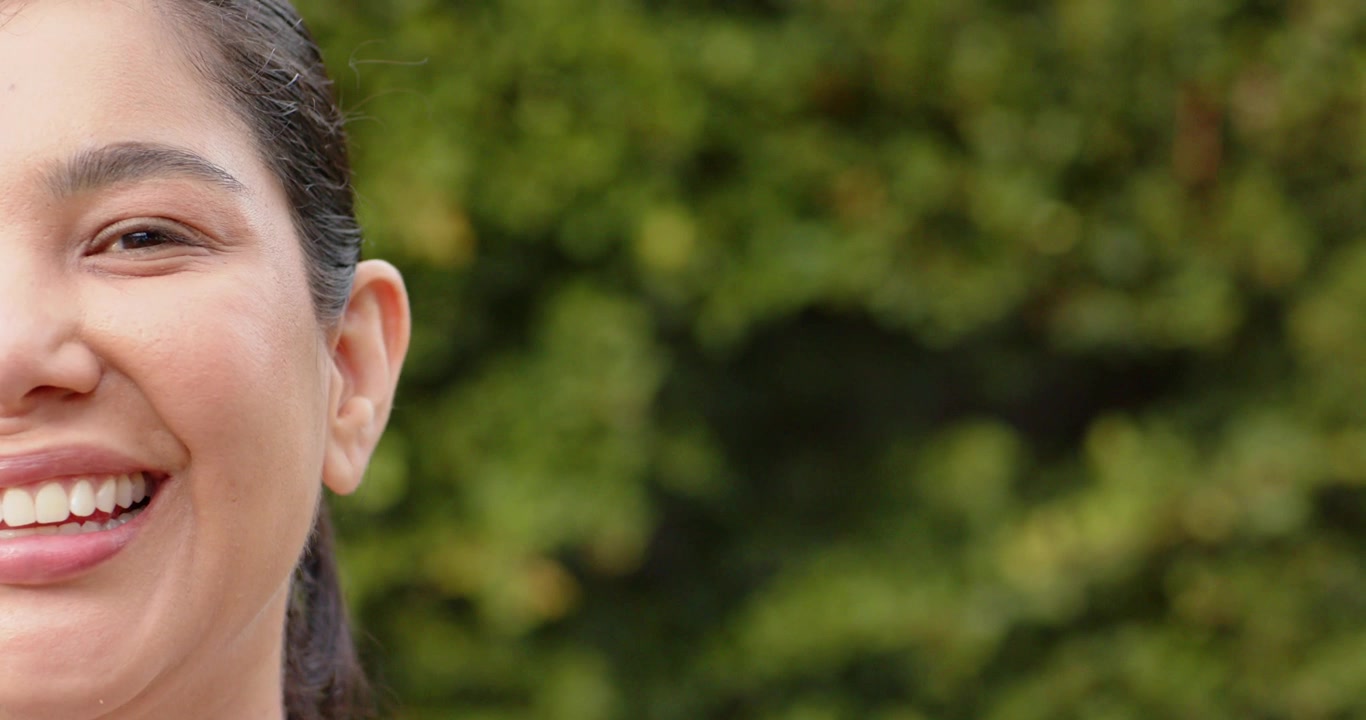 Donna di mezza età con i capelli legati, l'inquadratura che si stringe, il viso che si trasforma in un sorriso vicino a una siepe da giardino, spazio per il testo.