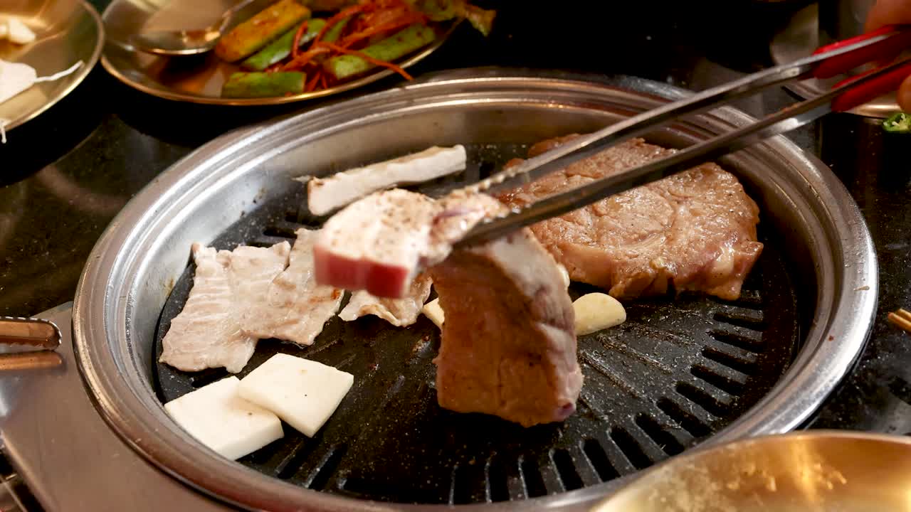 Hand uses tongs to place raw pork belly on hot Korean barbecue grill, overhead view
