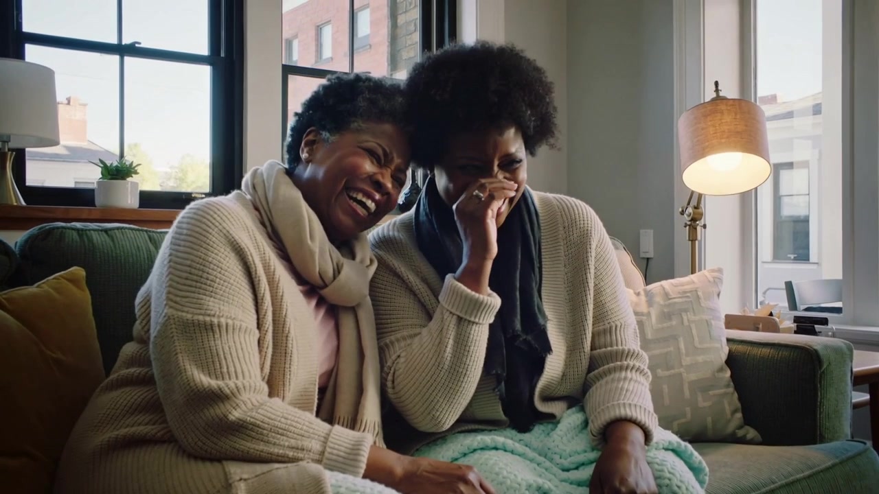 Due donne che ridono, indossando cardigan, mentre reagiscono a uno show televisivo a casa, condividendo popcorn su una coperta verde.
