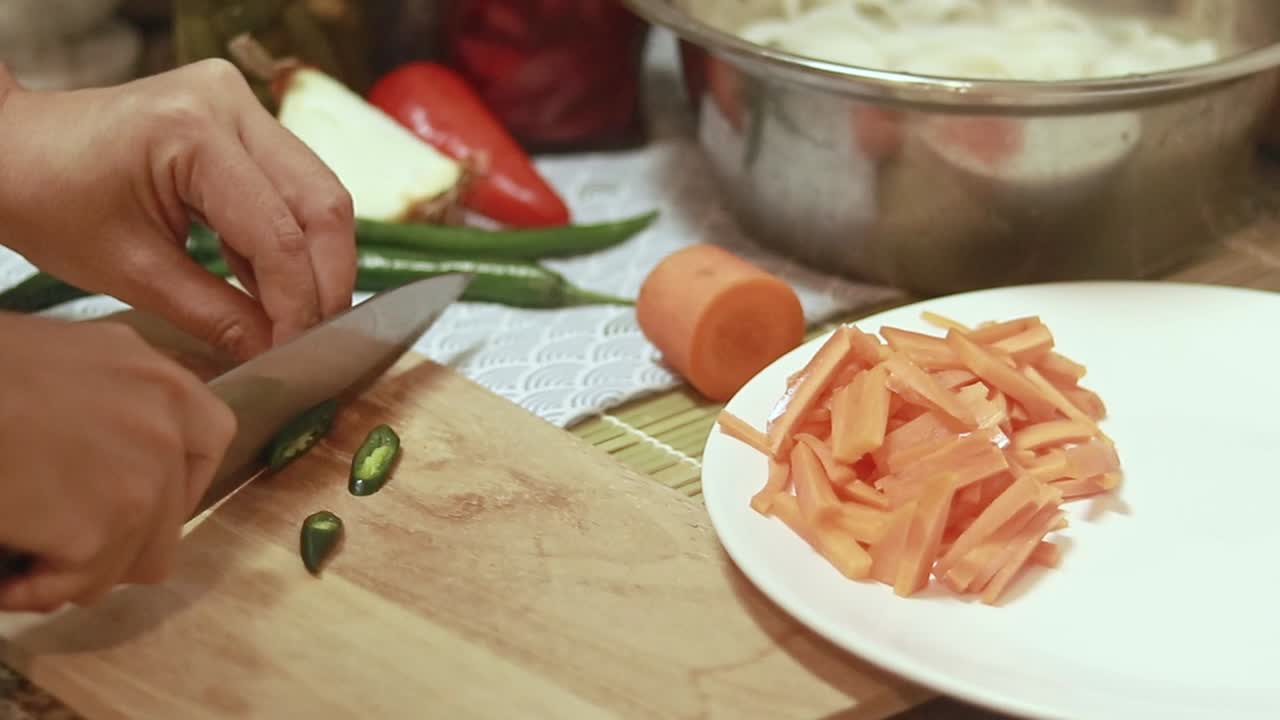 Hand slicing green chili into thin strips on a wooden board and setting it aside on a plate, preparing the ingredient for cooking at home