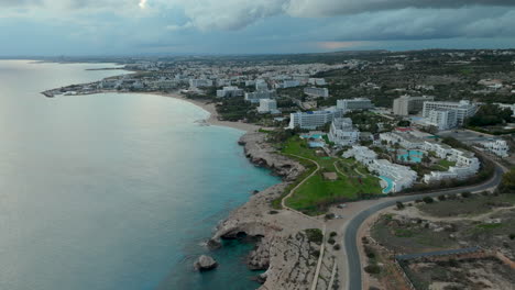 panoramic aerial view of ayia napa, expanse of the azure mediterranean sea alongside a curved coastline dotted with hotels and resorts, town stretching into the interior landscape under a moody sky