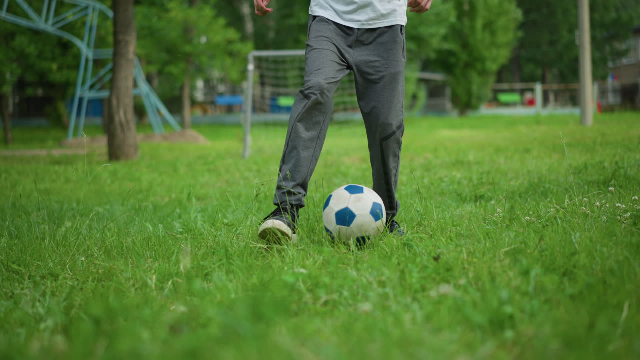un niño joven hábilmente dribla una pelota de fútbol en cámara lenta en un campo de hierba, el fondo revela una vista borrosa de un poste de la portería y el edificio