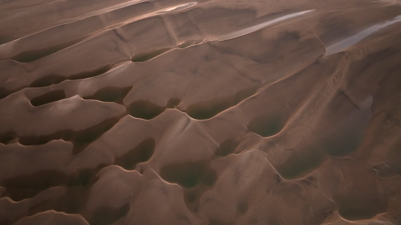 Intricate patterns of sand and seawater pools reflecting light at sunset. Forward flight over.